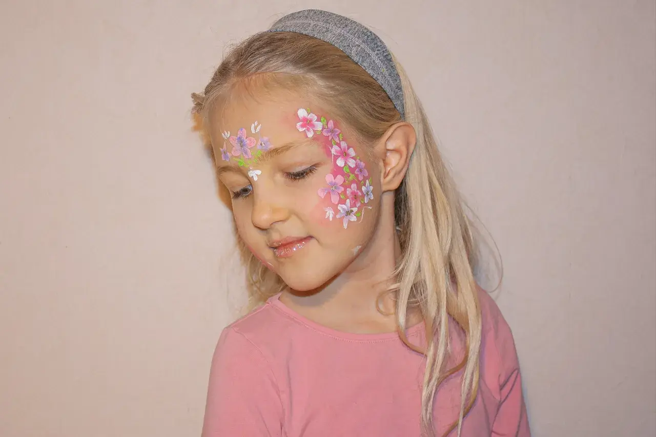 Ein Mädchen mit blonden Haaren und einem Stirnband, das ein Blumenmuster auf der Wange trägt. Das Stern schminken Kinder ist hier als zarte Blüten umgesetzt.