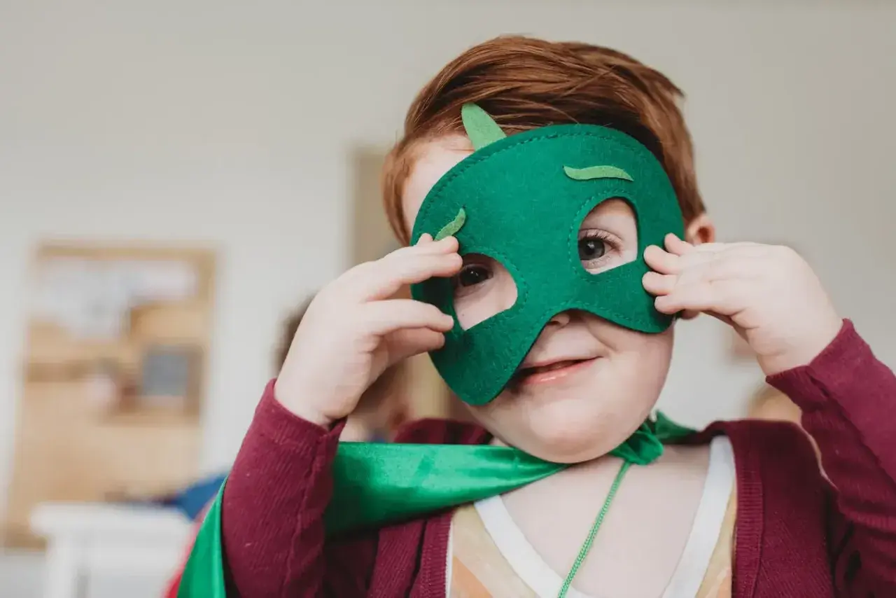 Ein Kind mit roten Haaren trägt eine grüne Drachenmaske und einen Umhang. Das Kind schaut neugierig durch die Augenlöcher.