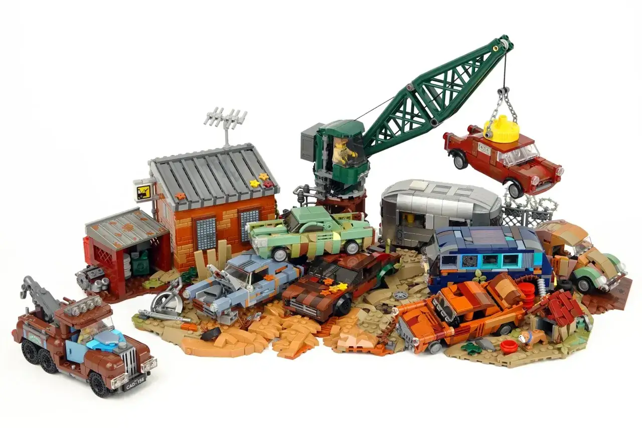 Ein detailreicher Lego Schrottplatz mit einem Kran, der ein Auto hebt, verschiedenen verrosteten Fahrzeugen, einem Wohnwagen und einem kleinen Gebäude.
