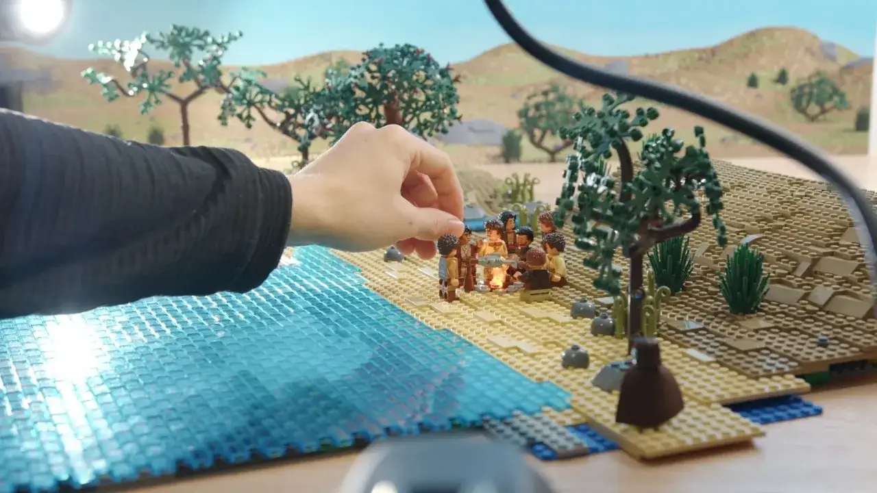 Eine Hand platziert Figuren f&uuml;r ein Lego Stop Motion am Strand. Eine Gruppe von Minifiguren versammelt sich um ein Lagerfeuer.