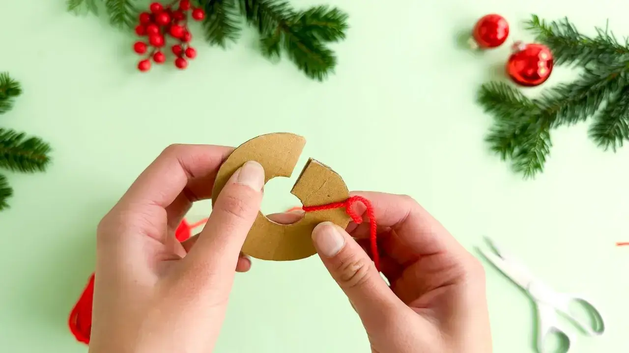 Hände wickeln roten Faden um Pappring für Basteln mit Pompons Weihnachten. Tannenzweige und Kugeln im Hintergrund.