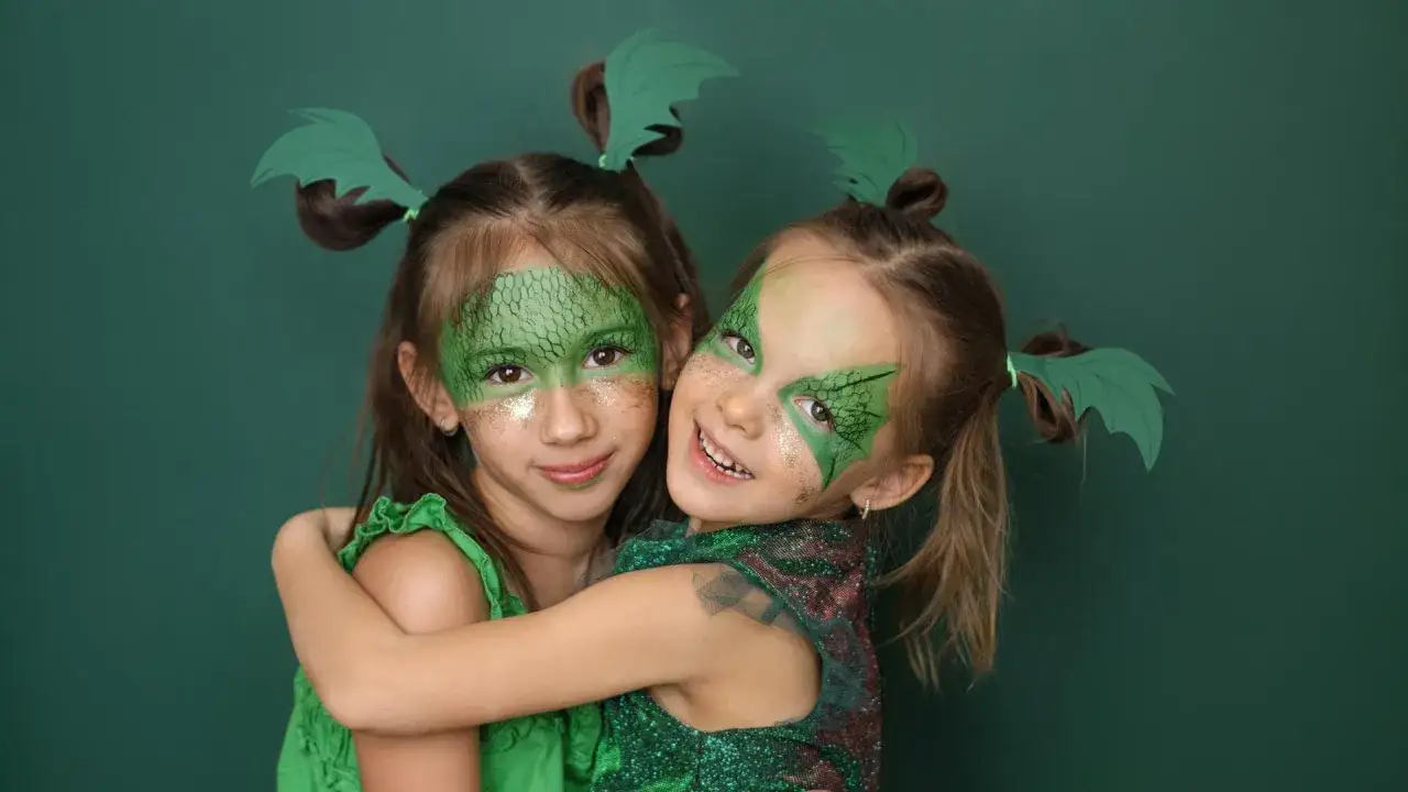 Zwei M&auml;dchen mit Drachen-Kinderschminken und gr&uuml;nen Fl&uuml;geln im Haar umarmen sich fr&ouml;hlich.