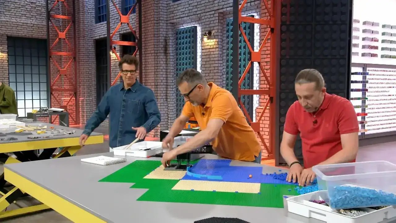 Drei Männer bauen mit bunten Lego-Steinen. Einer sortiert blaue Steine, ein anderer legt grüne und beige Platten aus. Ein dritter schaut zu. Hier sind die Lego Masters!