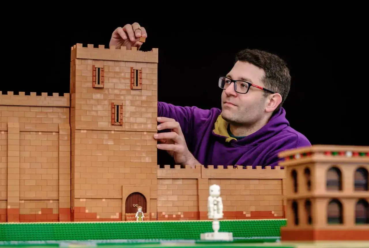 Ein Mann baut eine beeindruckende LEGO-Burg, ein wahres MOC (My Own Creation). Kleine Figuren und ein weiteres Gebäude sind bereits Teil der Szene.